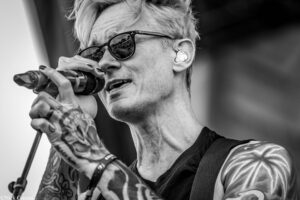 Male singer with tattoos wearing sunglasses, singing into a handheld microphone on a stage.