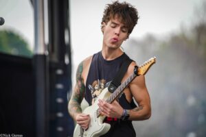 Guitarist with tattoos playing a white electric guitar on stage, wearing a sleeveless dark shirt and looking down at the fretboard.