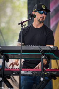 Male musician in a black t-shirt and cap playing a keyboard on stage, wearing blue mirrored sunglasses.