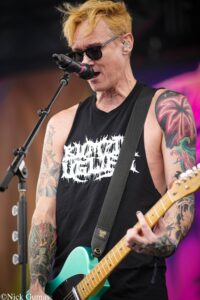 Male guitarist wearing sunglasses and a black sleeveless shirt, singing into a microphone while playing a turquoise electric guitar on stage.