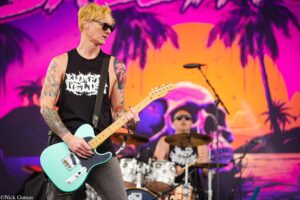 Guitarist in a black sleeveless shirt and sunglasses plays a turquoise electric guitar on a colorful stage backdrop with palm-tree design in pink and purple hues.