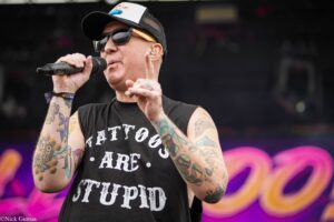 Tattooed male singer in a cap and sunglasses, singing into a handheld microphone on stage.