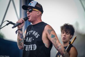 Male singer on stage singing into a microphone, wearing a cap, sunglasses, and a sleeveless shirt, with tattooed arms visible in the foreground.