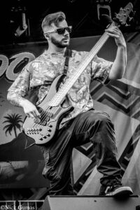 Male bass guitarist wearing sunglasses and a floral shirt, playing a five-string bass on stage during a live performance.