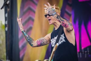 Tattooed guitarist with sunglasses on stage, arms raised and shouting during a live performance
