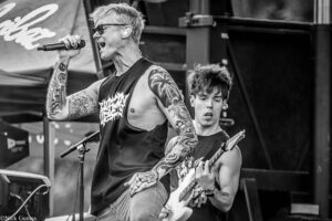 Male vocalist with tattoos and sunglasses sings into a microphone on stage, while a guitarist plays in the background (black-and-white photo).