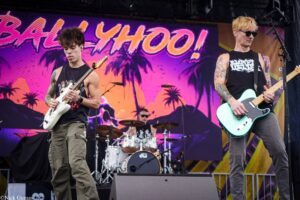 Two guitarists perform on stage with a tropical sunset backdrop; drummer visible in the background on a concert stage.