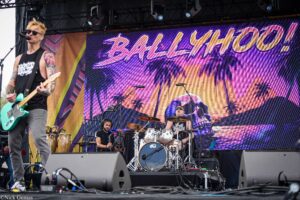 Band stage with guitarist left, turquoise-green guitar, tattoos, singing into mic; drummer behind drum kit on right; vibrant palm-tree backdrop.
