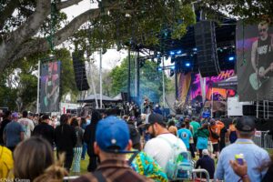 Outdoor concert stage with musicians performing, large screens, and a crowd in front.