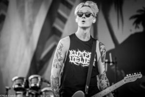 Guitarist on stage wearing sunglasses and a sleeveless shirt, heavily tattooed arms, playing a guitar in a black-and-white photo