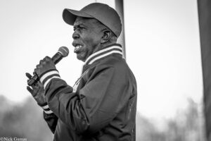 Black-and-white photo of a man wearing a cap and jacket, speaking into a handheld microphone outdoors.