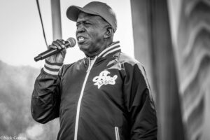 Black-and-white photo of a man in a cap singing into a handheld microphone outdoors.