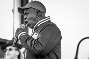 Black-and-white photo of a man singing into a microphone, wearing a cap and a striped-collar jacket, side profile.