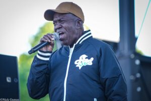 Man in a brown cap sings into a handheld microphone on an outdoor stage.