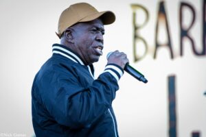 Man wearing a brown cap and navy jacket sings into a handheld microphone on stage, left side of the frame.