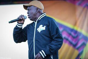 African American man wearing a tan cap and navy jacket sings into a microphone on stage with a colorful backdrop behind him.