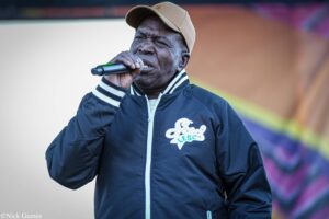 Man in a navy jacket and beige cap sings into a handheld microphone on stage.