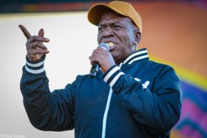 Male performer wearing a yellow cap and navy jacket sings into a handheld microphone on stage, gesturing with his other hand.