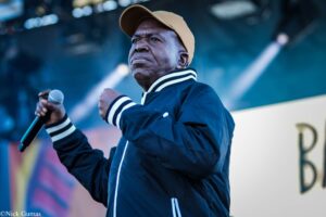 Singer on stage wearing a tan cap and navy jacket, gripping a microphone.