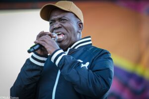 Male singer performing on stage, wearing a brown cap and navy jacket, holding a microphone close to his mouth.