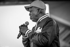 Side-profile of a man wearing a cap and varsity jacket, speaking into a handheld microphone at a microphone stand stage.