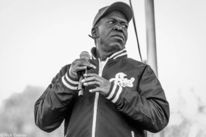 Black-and-white photo of a man wearing a baseball cap and jacket, speaking into a handheld microphone outdoors.