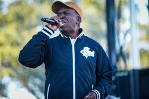 Man singing into a handheld microphone outdoors, wearing a navy jacket with white trim and a beige cap, against a blurred background.