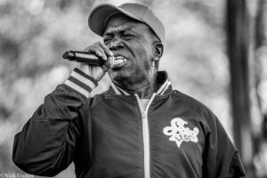 Black-and-white photo of a man singing into a handheld microphone outdoors, wearing a cap and jacket.