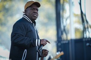 Older Black man in a tan cap and navy jacket on an outdoor stage, holding a microphone stand with a guitarist blurred in the background.