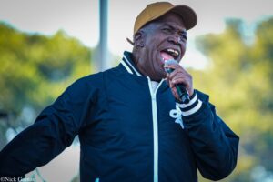 Man wearing a brown cap and dark jacket singing into a microphone outdoors with a blurred green background.