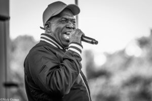 Black-and-white photo of a man singing into a handheld microphone on stage, wearing a cap and striped-hem jacket outdoors.