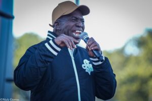 Man wearing a tan cap and black jacket sings into a microphone at an outdoor event, smiling widely.