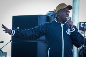 African American man wearing a beige cap and navy jacket, singing into a microphone on a stage with a large speaker behind him outdoors
