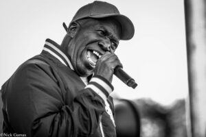 Black-and-white photo of a man wearing a cap and jacket, singing into a handheld microphone on stage.