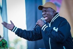 Man wearing a tan baseball cap and navy jacket sings into a handheld microphone on stage, arm extended toward the audience.