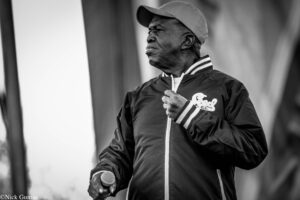 Black-and-white photo of a man wearing a cap and jacket, holding a microphone as if performing on stage.
