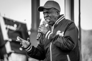 Male performer singing into a handheld microphone, wearing a cap and jacket, with an expressive facial moment on stage.