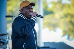 Man singing into a handheld microphone outdoors, wearing a tan cap and navy jacket with white striped cuffs.