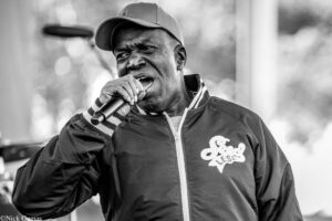 Black-and-white image of a male singer wearing a cap and jacket, holding a microphone and performing closely to the camera.