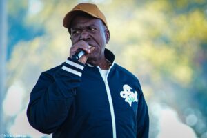 Man wearing a tan cap and navy jacket sings into a handheld microphone outdoors, posed close to the camera.