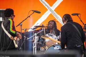 Drummer with dreadlocks playing a drum kit on stage, flanked by two guitarists against an orange backdrop with white X shapes.