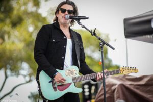 Male guitarist performing on an outdoor stage, wearing sunglasses and a black jacket, playing a turquoise guitar into a microphone