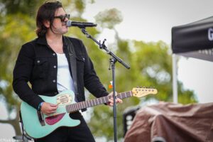 Male musician in a black jacket and sunglasses plays a turquoise electric guitar and sings into a microphone outdoors.