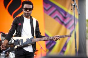 Musician wearing sunglasses and a black jacket with colorful stripes plays a bass guitar on a brightly painted stage backdrop.