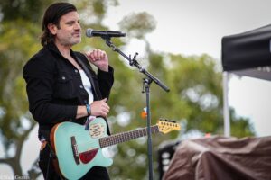 Male musician singing into a microphone while playing a turquoise guitar outdoors on stage.