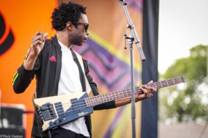 Male musician wearing a black Adidas jacket and sunglasses, playing a bass guitar on an outdoor stage with an orange backdrop.