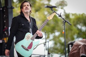 Male guitarist on stage playing a turquoise electric guitar with a sunburst pickguard, wearing a black shirt, in an outdoor concert setting.
