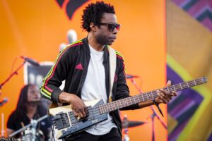 Male bass guitarist wearing sunglasses and a black Adidas jacket with neon stripes, playing a bass on a brightly colored stage backdrop.