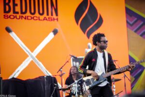 Musician playing a bass on stage with an orange backdrop and flame logo behind him.