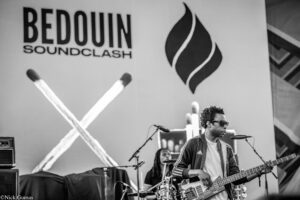 Stage performance featuring a bassist in sunglasses playing a bass guitar, with a Bedouin Soundclash backdrop in black and white.